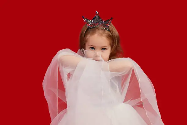 Joyful little girl with long hair in a tulle skirt and princess sparkling crown on her head isolated on red background. Celebrating a colorful carnival for kids, expressing positive birthday