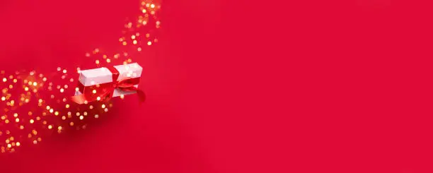 Photo of Big gift box wrapped in wrapping pink paper and a red satin ribbon bow flying in the air on a red background with copy space.