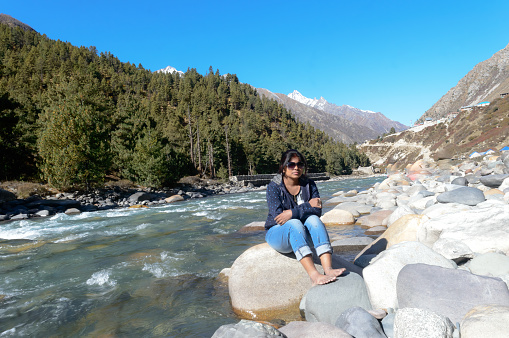 Front View of Solo Indian traveler in winter casual sitting alone near stream flowing water of remote mountain valley. Snow capped Himalayan mountain forest blue sky in background. Spiti Valley Himachal Pradesh India South Asia Pac