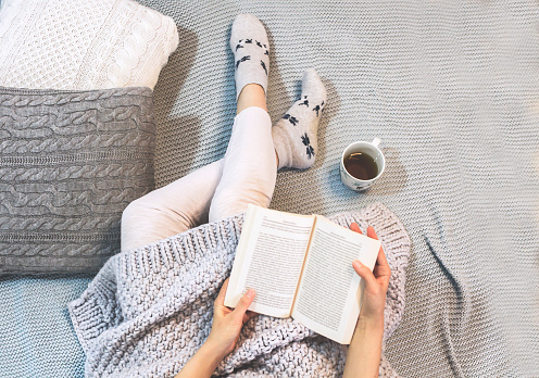 Young woman sitting on a bed, holding a book, beside her is a cup of hot tea, winter scene