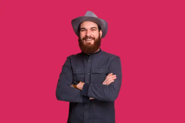 Handsome bearded cowboy with foldedhands looking at camera standing over isolated background