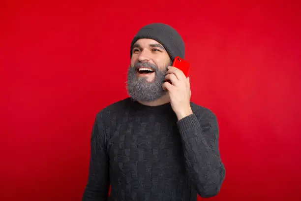 Happy bearded hipster man standing over red background and talking on smartphone