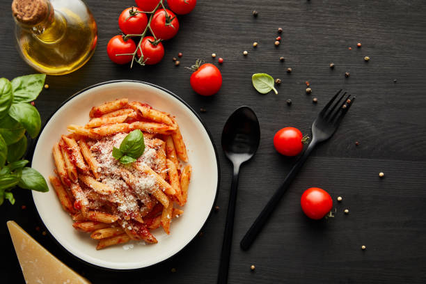 top view of tasty bolognese pasta with tomato sauce and parmesan in white plate near ingredients and cutlery on black wooden background - penne imagens e fotografias de stock