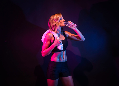 Waist up / one person of 20-29 years old adult beautiful caucasian young women / female standing in front of black background / multi-colored background / colored background wearing sports clothing / sports bra / shorts / running shorts who is drinking and holding or wearing a towel and holding water bottle / black color / gel effect lighting