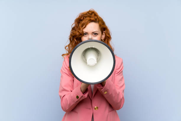 istock Redhead woman in suit over isolated blue wall shouting through a megaphone 1189403968