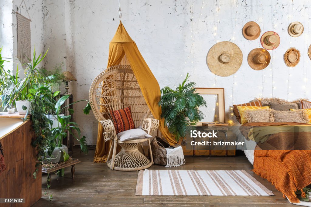 Cozy house with room in boho style interior Elegant and quiet bohemian room with cozy interior, wicker chair, pillows, cushions, green plants in flower pot, bed and rug on wooden floor Boho Stock Photo