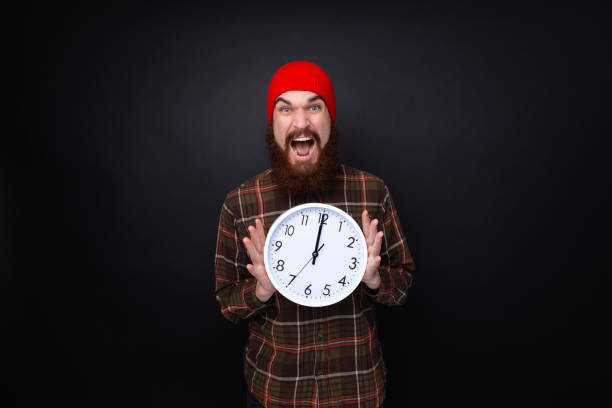 istock Excited bearded guy in red hat, holding a clock standing over dark wall background 1189418063