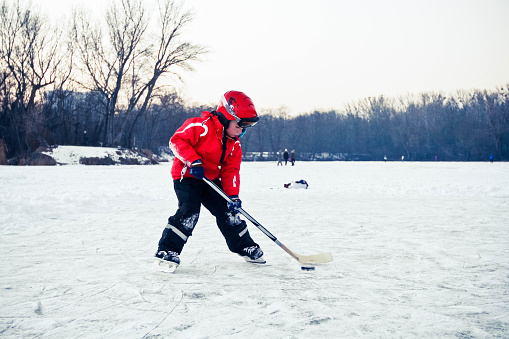 A boy in a helmet with ski goggles, dressed in red and black winter clothes is holding a hockey stick. He is concentrating on the puck.