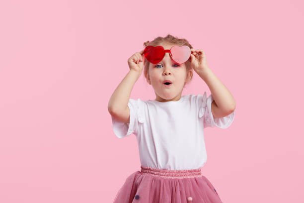 porträt von überrascht niedlichen kleinen kleinkind mädchen in der herzform sonnenbrille. kind mit offenem mund mit spaß isoliert über rosa hintergrund. blick auf die kamera. wow lustiges gesicht - child glasses eyewear little girls stock-fotos und bilder