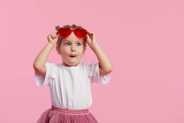 ハート型サングラスで驚いたかわいい幼児の女の子の肖像画。ピンクの背景に隔離された楽しみを持つ開いた口を持つ子供。カメラを見て。うわー面白い顔 - child surprise little girls human face ストックフォトと画像