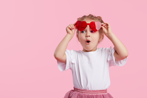 Retrato de una niña pequeña dazalda sorprendida en las gafas de sol en forma de corazón. Niño con la boca abierta divirtiéndose aislado sobre fondo rosa. Mirando la cámara. Cara divertida Wow photo