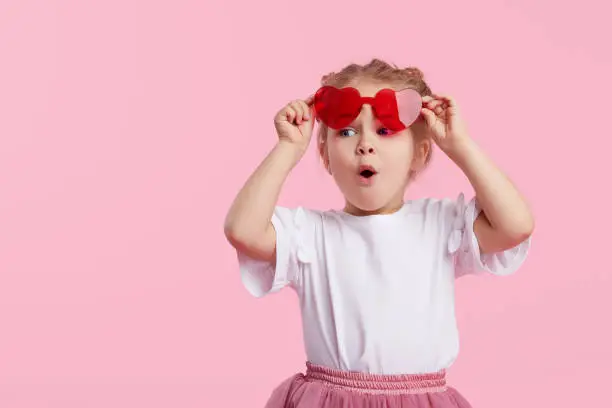 Photo of Portrait of surprised cute little toddler girl in the heart shape sunglasses. Child with open mouth having fun isolated over pink background. Looking at camera. Wow funny face