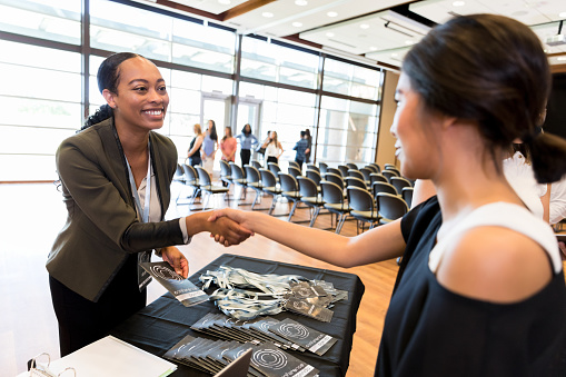 Cheerful mid adult businesswoman shakes hands with a colleague during a conference. The businesswoman is working at the conference registration table.