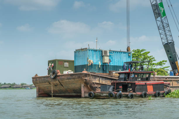 Tan Phong, Mekong Delta, Vietnam - March 13, 2019: Brown Pontoon, green crane, blue containers and red tugboat at working site along shore under blue cloudscape.
