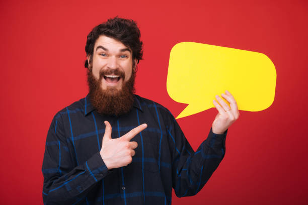 istock Portrait of a cheerful bearded guy holding empty  speech bubble and pointing finger over red background 1189423325