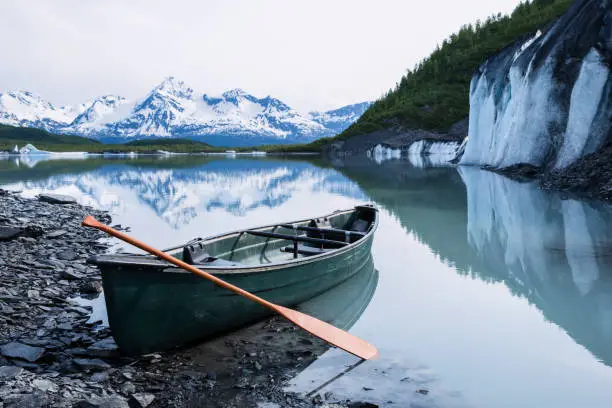 Canoe resting on the edge of Valdez Glacier, covered in rock and till (moraine) on the lower glacier above Valdez Lake. Large ice cliff of the glacier and icebergs can be seen beyond.