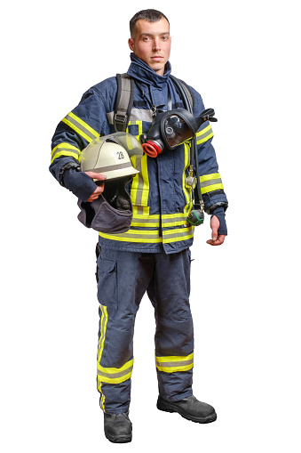 A young brave fireman in a fireproof uniform stands and looks at the camera with a helmet in his hands. Isolated on white