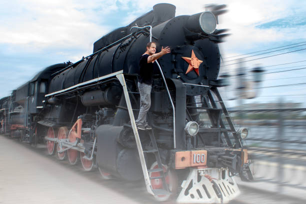 istock Arrival of old train at the station. Technique since second world war. Retro locomotive pulls up to platform. A man stands on a vintage steam locomotive. Vintage style on a cloudy sky background. 1189444953