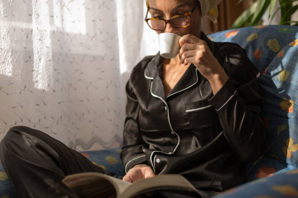 Beautiful woman at her home read a book and drink a cup of coffee sitting in sofa against a white curtained windows wearing pajamas