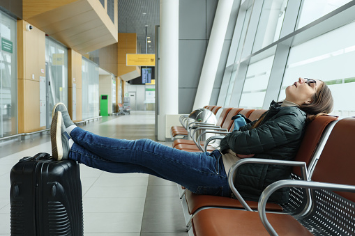 Bored and tired young woman is waiting her flight in airport sitting in hall. She sits and sleeping with her legs stretched out and puts them on the suitcase.