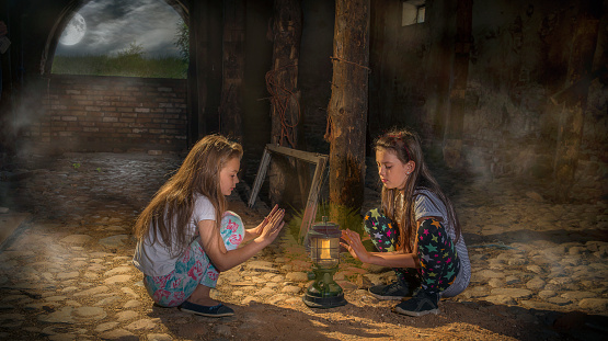 Girls in an old,abandoned building sit near a lamp and warm themselves.The moon is visible from the window through the fog.