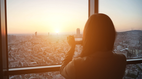Silhouette of woman taking a pictures of panoramic city view at sunset on smartphone, focus on the city