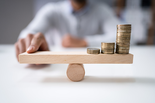 Close-up Of A Businessman Balancing The Coin Stack On Wooden Seesaw Over Desk