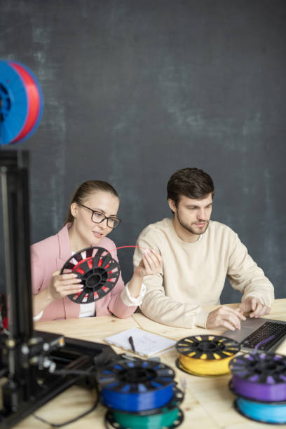 istock Young female looking at red filament on spool while sitting next to colleague 1189510353