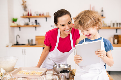 Adorable joyful child with touchpad showing his mom video recipe of something really yummy while choosing what to cook