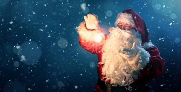Photo of Happy Santa Claus holding glowing christmas bauble over defocused blue background