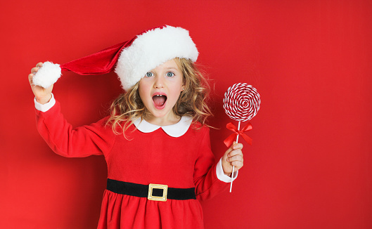 Surprised christmas girl in a Santa hat and red dress holding a candy. Happy Christmas and New Year. Concept of celebrating New year and Christmas.