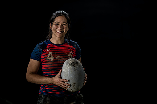 Portrait of smiling female rugby player holding ball against black background.