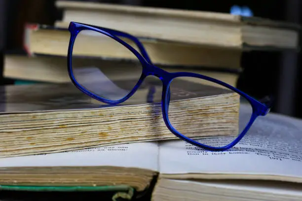 Photo of View on isolated stack of old yellowed books on wood table with blue reading glasses and open book