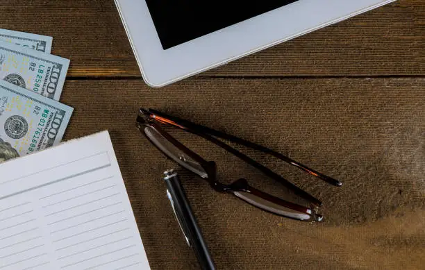 Photo of US currency money in white table touch screen on notepad, pen and glasses workspace desk with wooden table