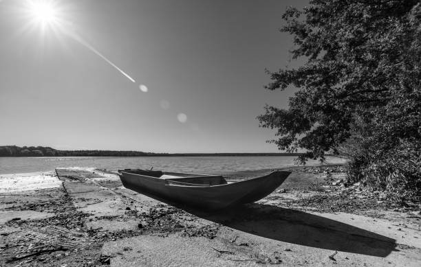 Natural still life. Fishing dinghy on shore, concrete pier, tree, rippled water surface and sunbeams on sky above horizon. Pisciculture. Horusicky fishpond in South Bohemia