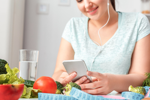 Young chubby woman wearing earphones standing in kitchen sitting at table holding smartphone listening to music close-up smiling joyful blurred background