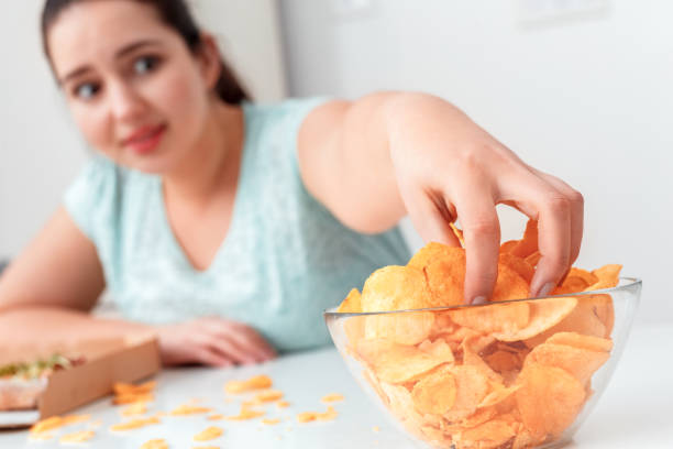 istock Breaking Diet. Chubby girl sitting at kitchen table eating chips excited close-up blurred background 1189616523