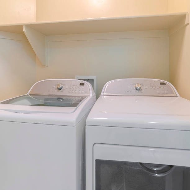 istock Square Washing machine and dryer inside a laundry room with shelf and beige wall 1189642873