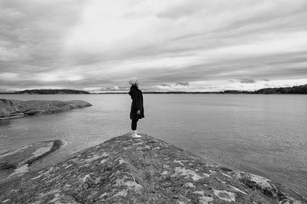 Black and White background image of woman wearing winter and raincoat boots and hat standing on thriangle Island rock on the sea looking up to sky, feel grey Lonely sadness winter concept.