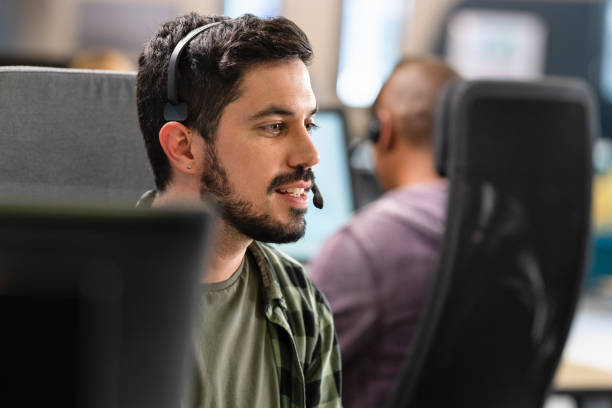 Man Talking To A Customer Over A Headset Hispanic man using a computer and talking to a customer over a headset while doing customer support in a call centre. iberian ethnicity stock pictures, royalty-free photos & images