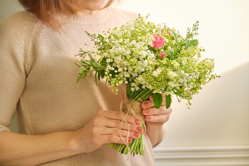 Beautiful fresh bouquet of lily of the valley flowers, pink rose, green branches in woman hand, background light sunny wall. Floristry, work of florist, flower shop, beauty