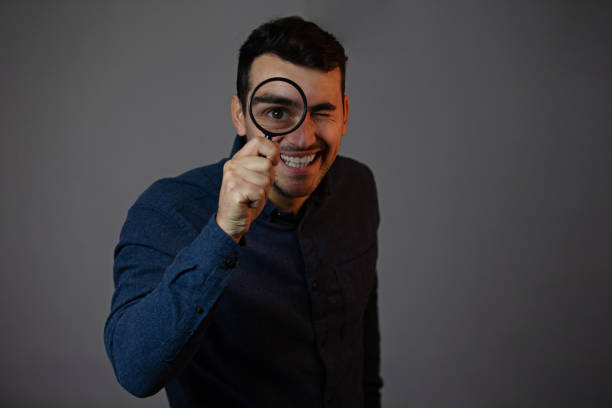 灰色の背景に虫眼鏡を持つ笑顔の男。灰色の背景に隔離された虫眼鏡を持つ好奇心旺盛な若者。 - magnifying glass lens holding europe ストックフォトと画像
