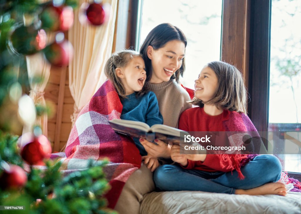 mother reading to daughters near Christmas tree. Merry Christmas! Pretty young mother reading a book to her daughters near Christmas tree. Christmas Stock Photo