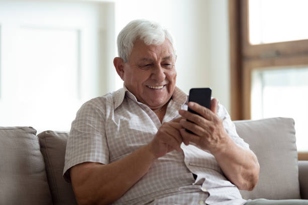 hombre mayor feliz sentado en el sofá en casa, usando el teléfono inteligente. - medical texts fotografías e imágenes de stock