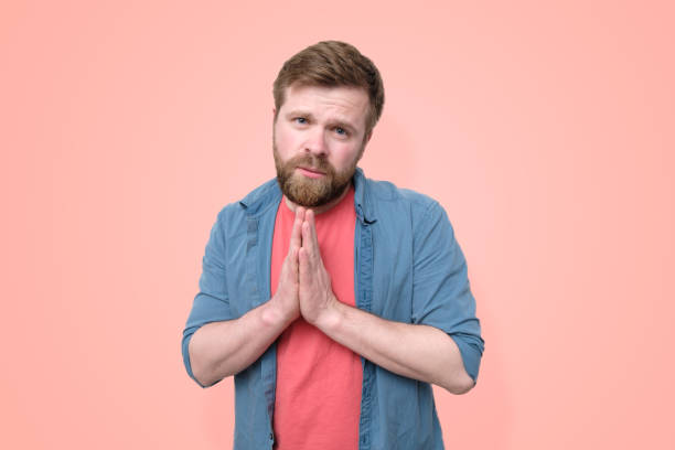 istock A young, Caucasian, bearded man with an imploring expression cupped his palms together and looked at the camera pitifully. Isolated on a coral background. Trendy colors. 1189754871