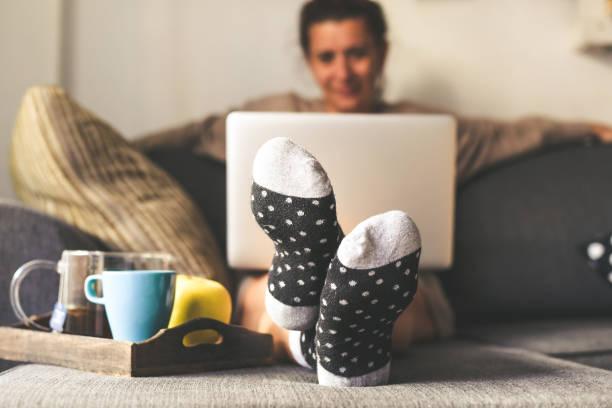 woman sitting on a couch in the living room with warm socks in a winter morning. girl using laptop and works at home, having natural breakfast with tea coffee and fruits. focus on feet in foreground - sensuality lifestyles cheerful comfortable imagens e fotografias de stock