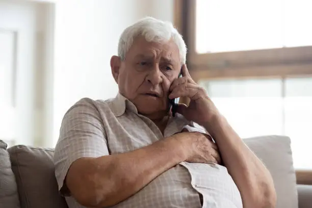 Worried older unhealthy man sitting on couch, making emergency 911 call, having painful feelings in chest, heart attack disease symptoms. Unhappy frustrated elderly grandfather listening to bad news.