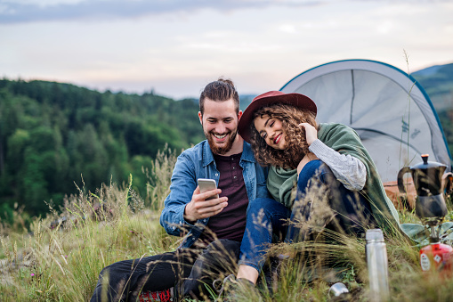 Young tourist couple travellers with tent shelter sitting in nature in the evening, using smartphone.