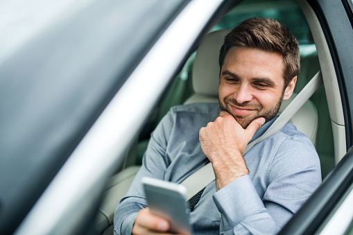 Young businessman with shirt, tie and smartphone sitting in car, text messaging.