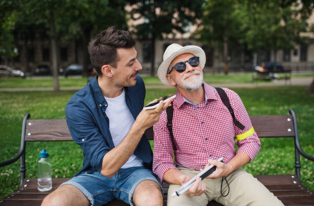 Young man and blind senior sitting on bench in park in city, using smartphone. A young man and blind senior sitting on bench in park in city, using smartphone. aids walk stock pictures, royalty-free photos & images